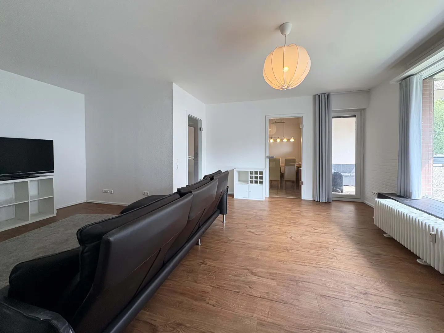Bright living room with wood floors, white walls, and a black leather sofa. A dining room is visible through a doorway.