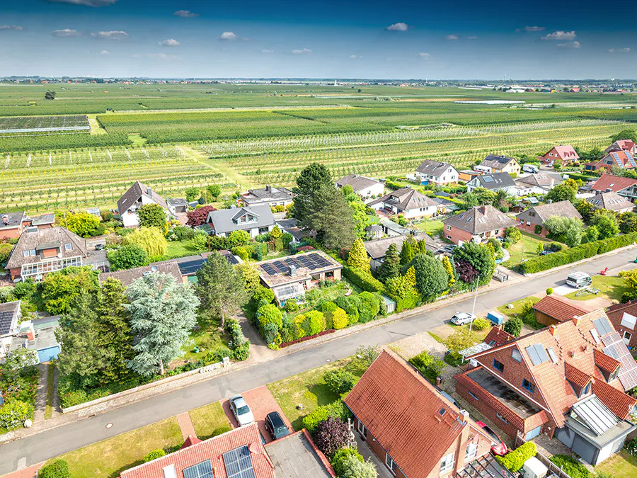 Aerial view of a suburban neighborhood with red-roofed houses, lush green trees, and vast agricultural fields beyond.