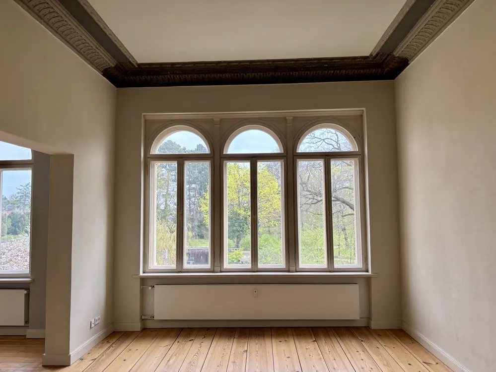 Empty room with wood floors, cream walls, and a large window with three arched panes overlooking trees. Ornate ceiling trim.