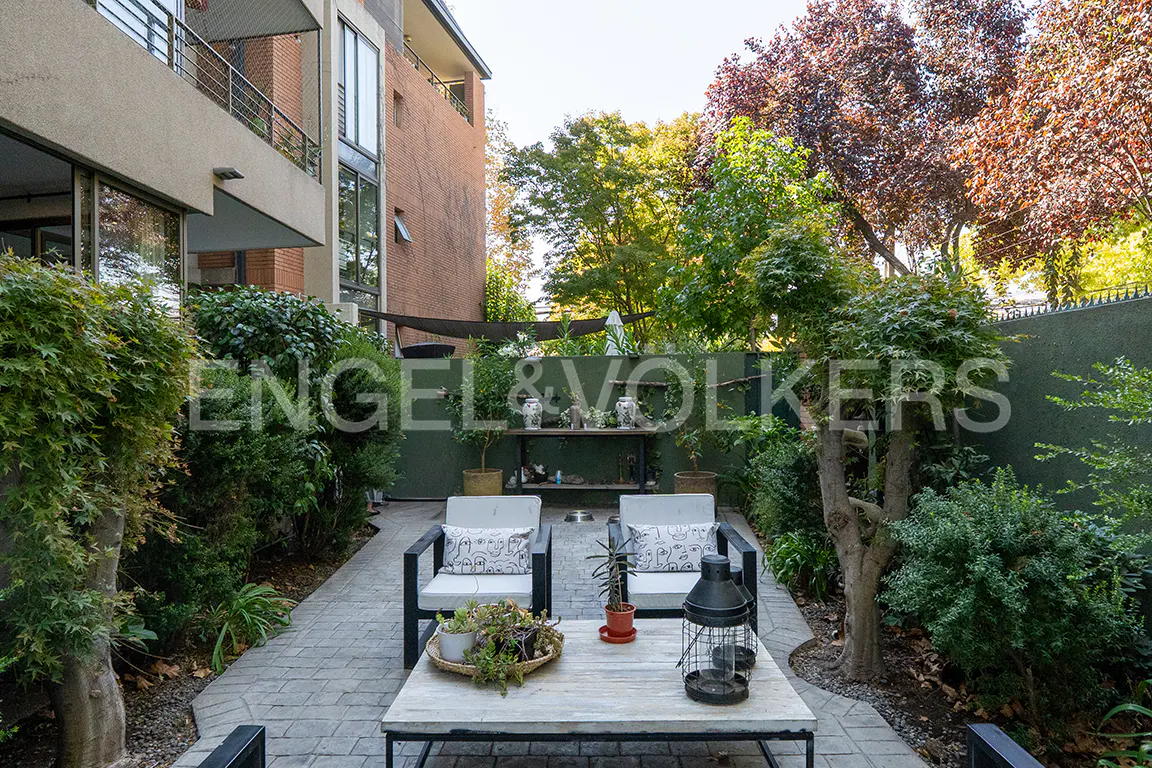 Outdoor patio with two chairs, a table, and greenery. The patio is paved with gray stone and surrounded by trees and bushes.