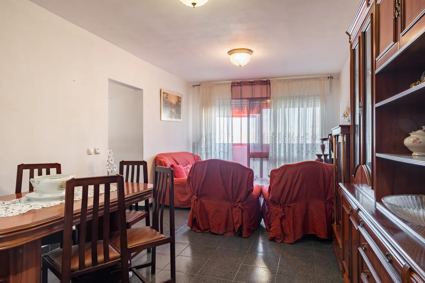 A living room with a dark wood dining table, red sofa and armchairs, and a large dark wood china cabinet.