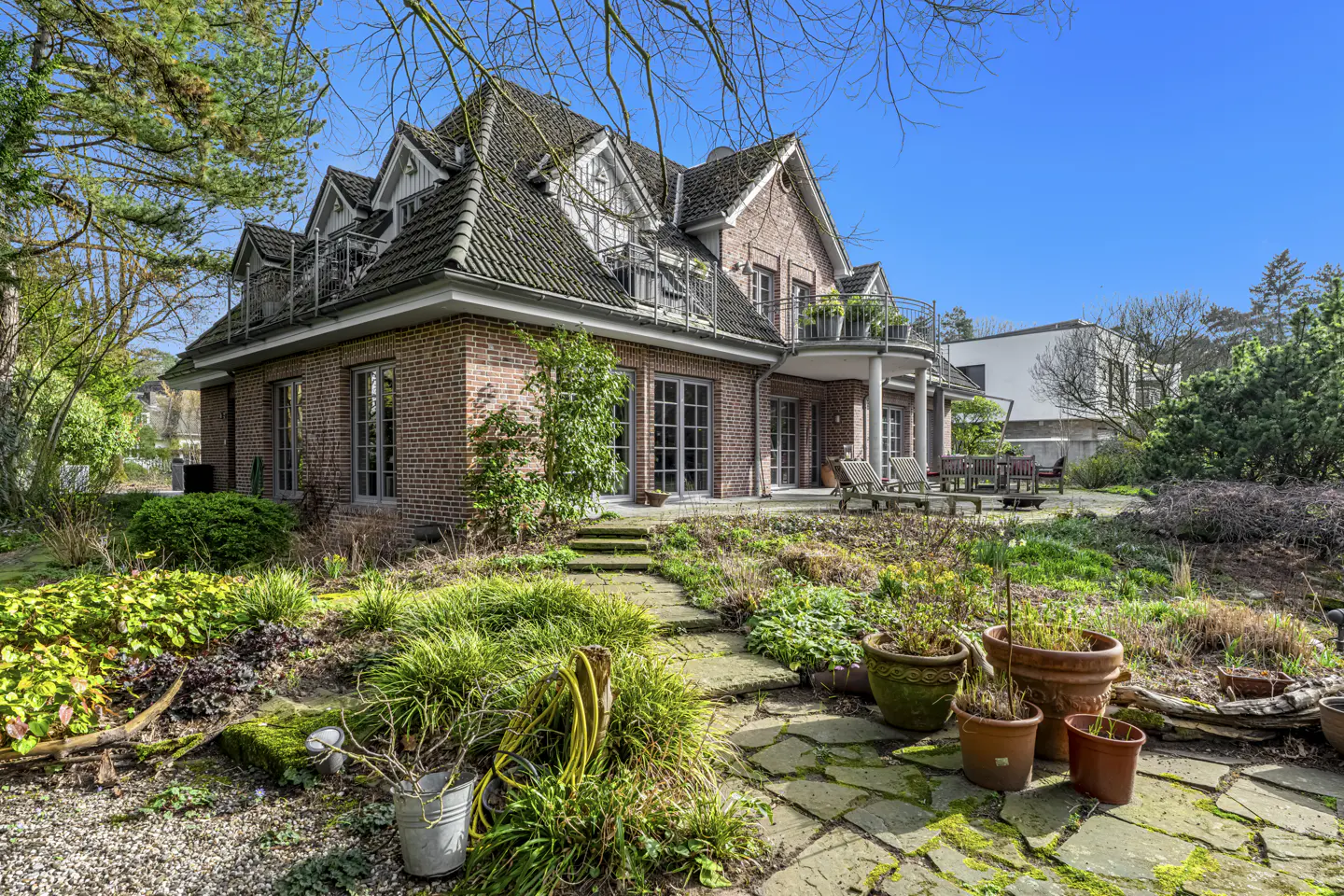 Brick house with a gray tiled roof, dormers, and balconies, surrounded by a lush green garden with stone pathways and potted plants.