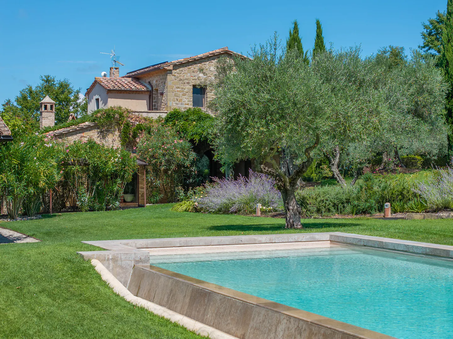 A stone house with a red tile roof overlooks a turquoise pool surrounded by green grass and trees.