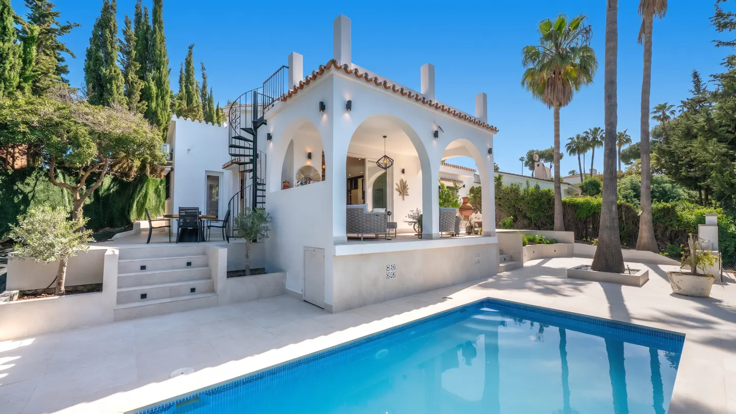 Exterior of a white villa with a pool, arches, and a spiral staircase under a clear blue sky. Palm trees and greenery surround the property.