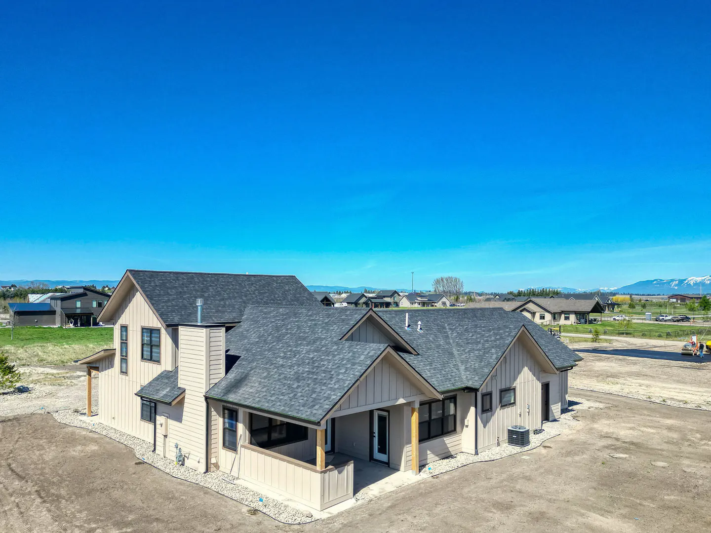 Beige house with a gray roof under a blue sky. The house has a covered porch and is surrounded by gravel. Other houses are in the background.