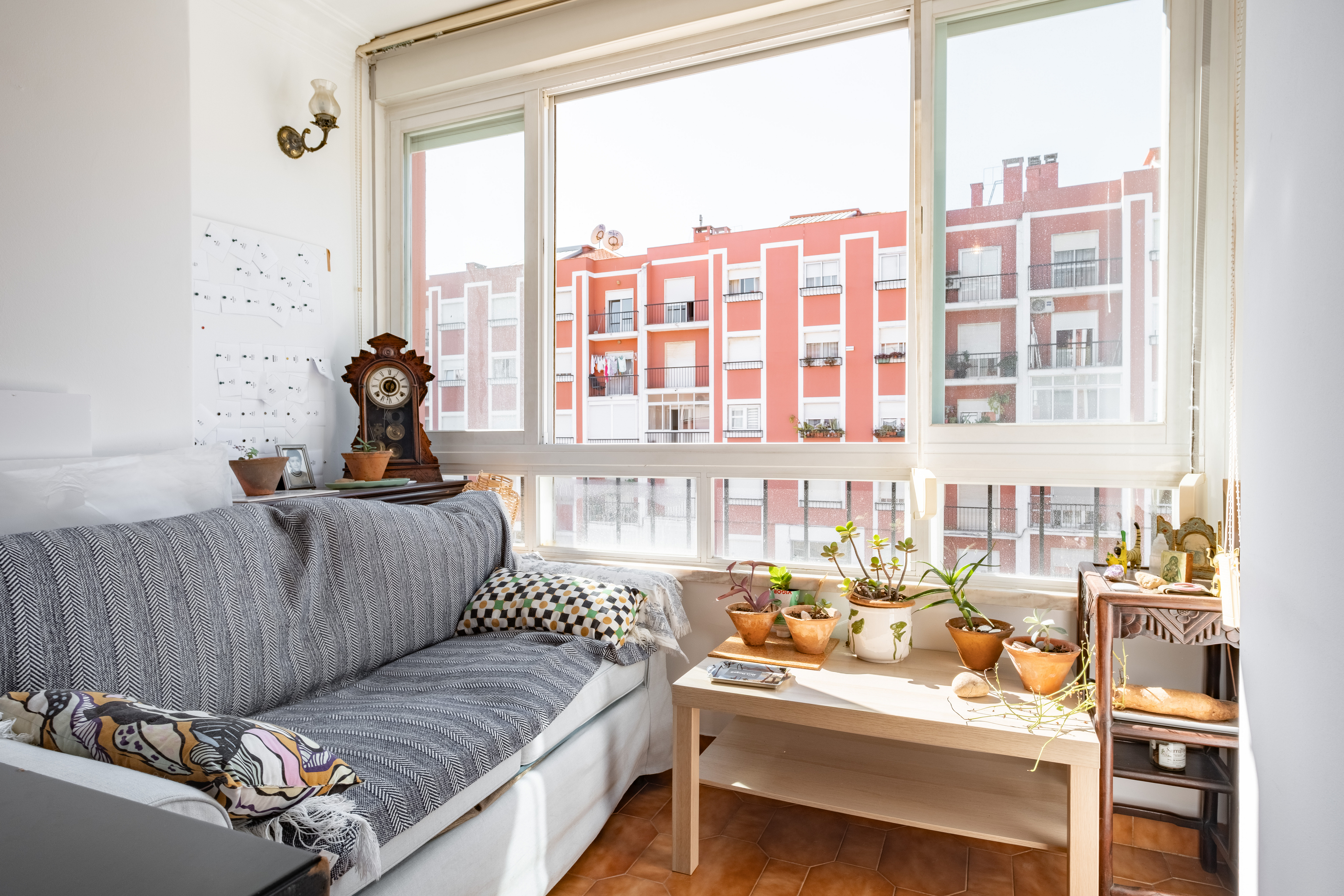 Sunlit sunroom with a grey sofa, wooden coffee table with potted plants, antique clock, and view of red apartment buildings. [AI-generated text]