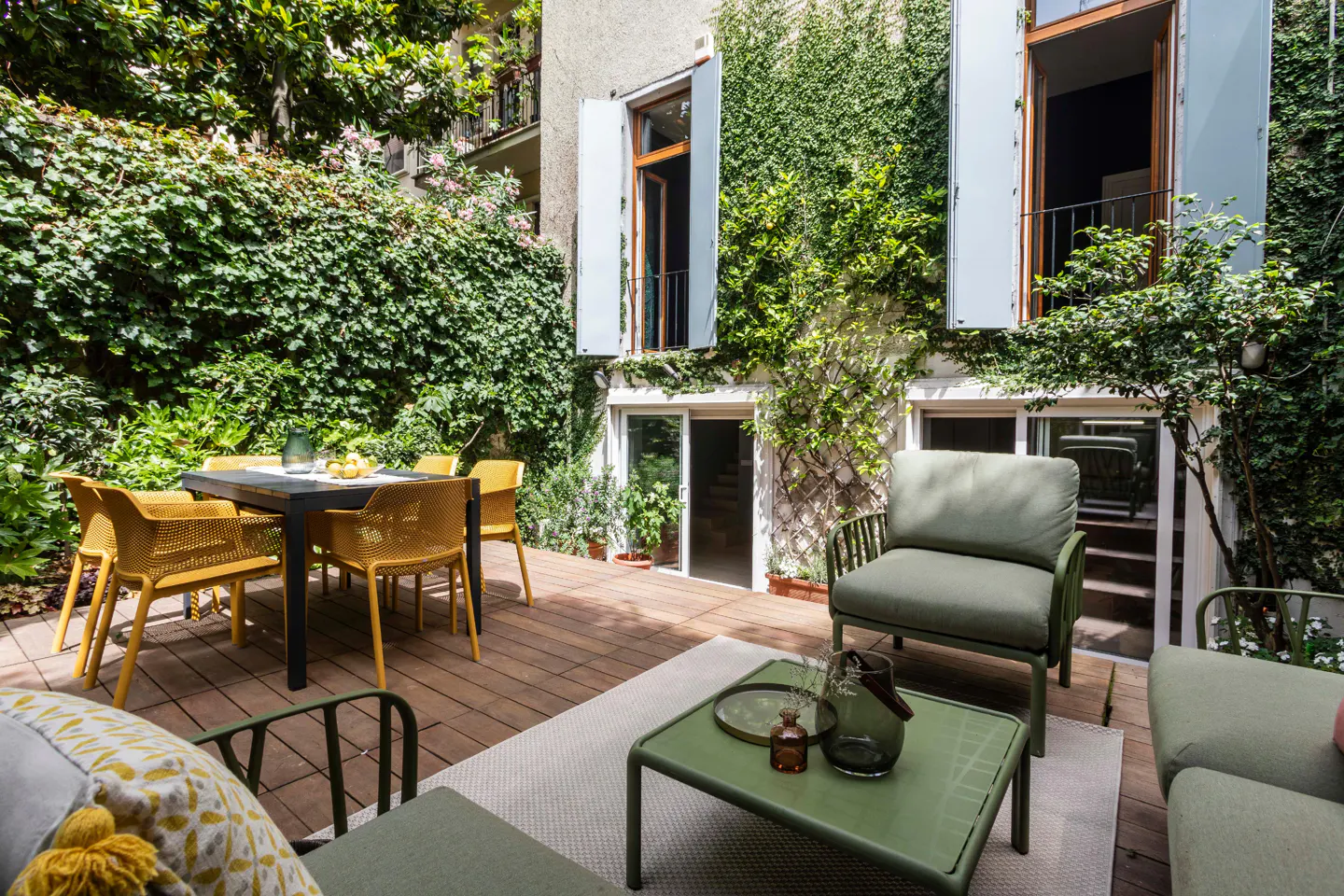 Outdoor patio with yellow chairs around a black table, green seating area, and building covered in ivy.