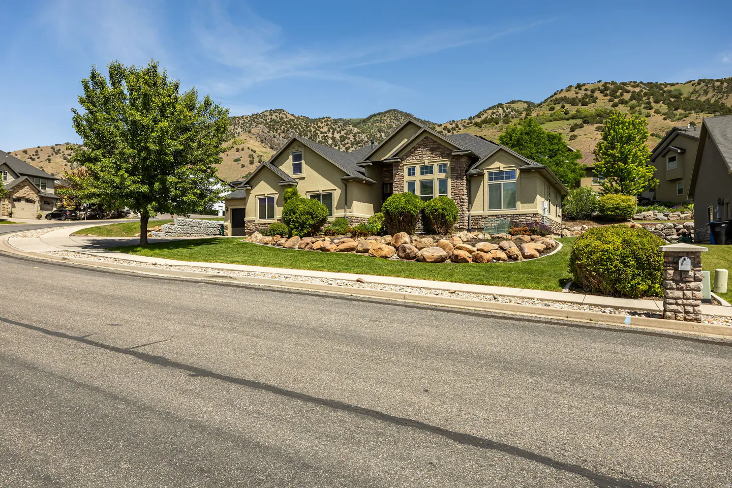 Tan suburban home with stone accents, manicured lawn, and large boulders, set against a backdrop of rolling mountains.