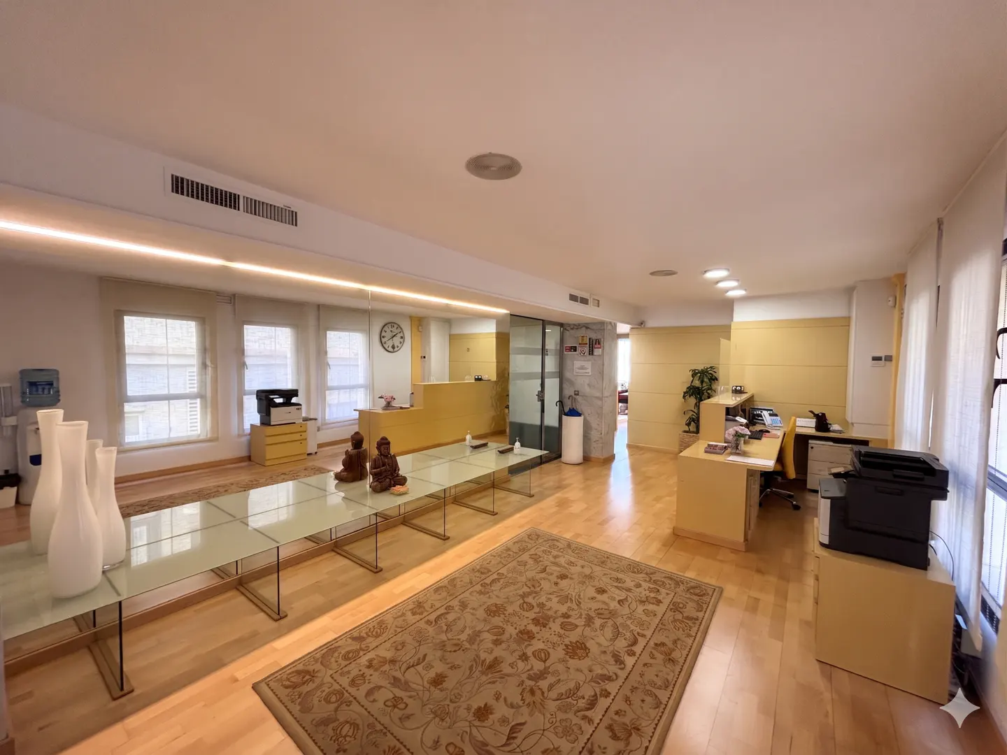 Bright office space with wood floors, a long glass table, and a reception area with yellow desks. Vases and Buddha statues decorate the table.