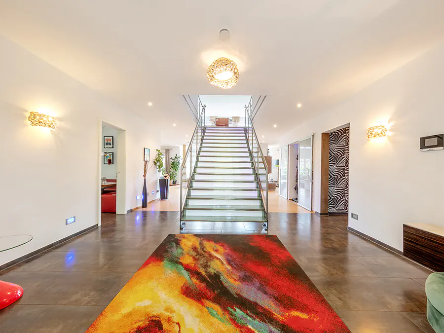 Bright foyer with white walls, brown tile floor, and a colorful rug. A staircase with metal railings leads to the upper level. Decorative lighting fixtures adorn the walls.