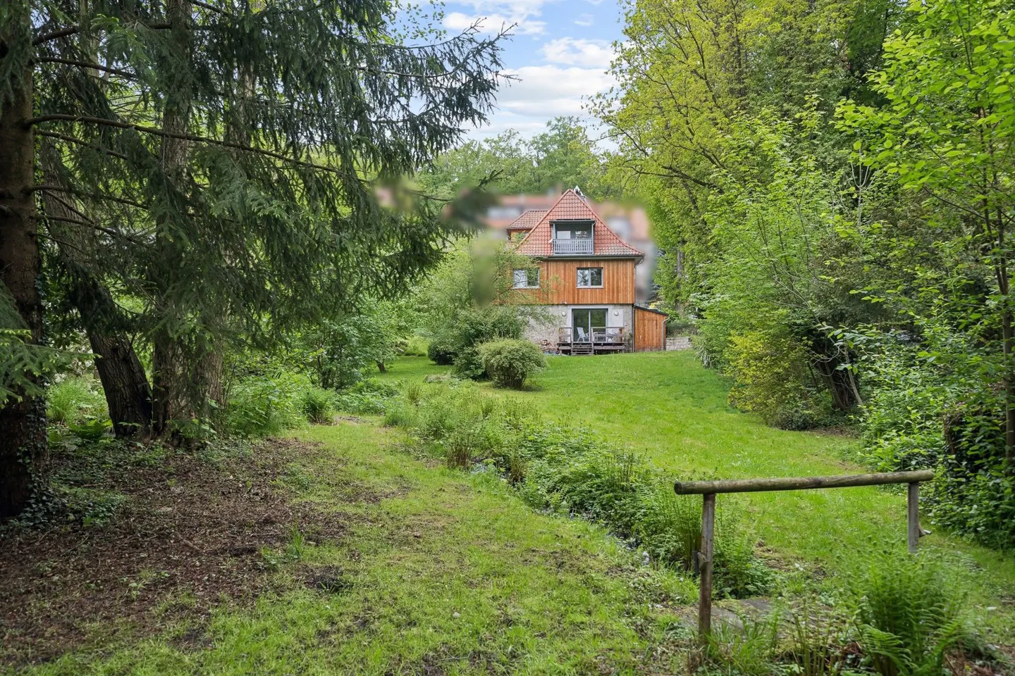 View of a wood-sided house with a red roof, seen through a green, wooded area with a grassy path.