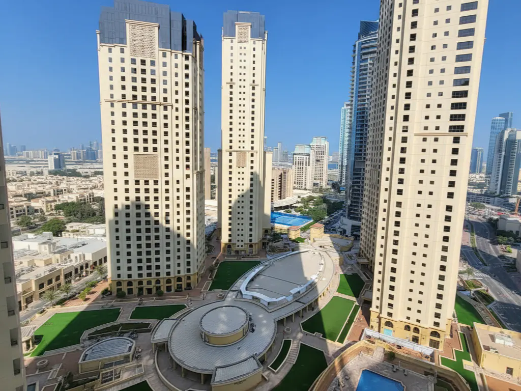 View of Dubai Marina with tall beige buildings, green spaces, and a swimming pool under a clear blue sky.