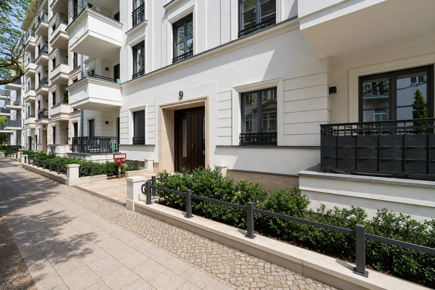 Exterior view of a white apartment building with dark windows and balconies, a brown door, and a stone walkway.