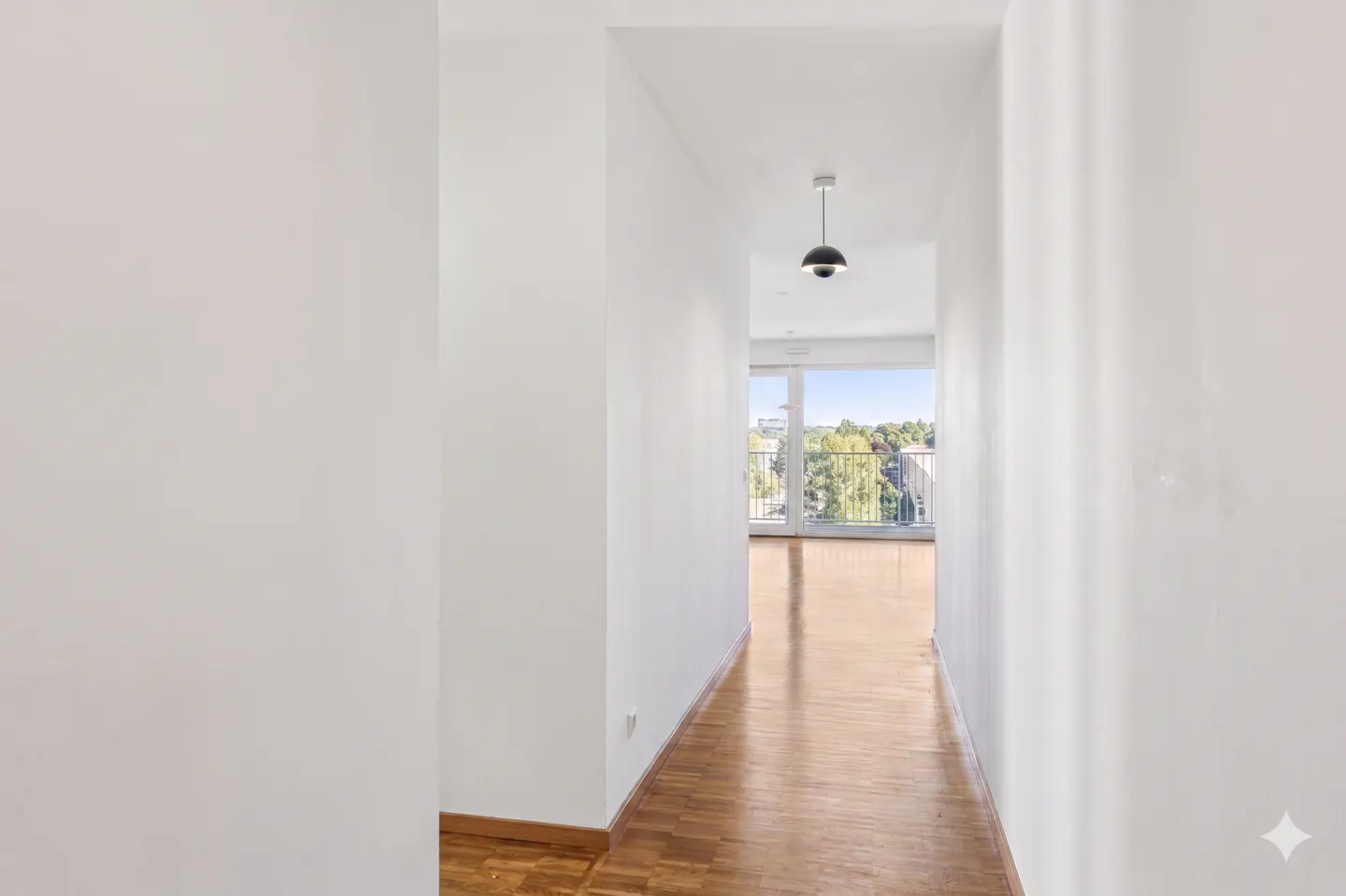Hallway with white walls and parquet floor leading to a bright room with a balcony and view of trees. A black pendant light hangs in the hallway.