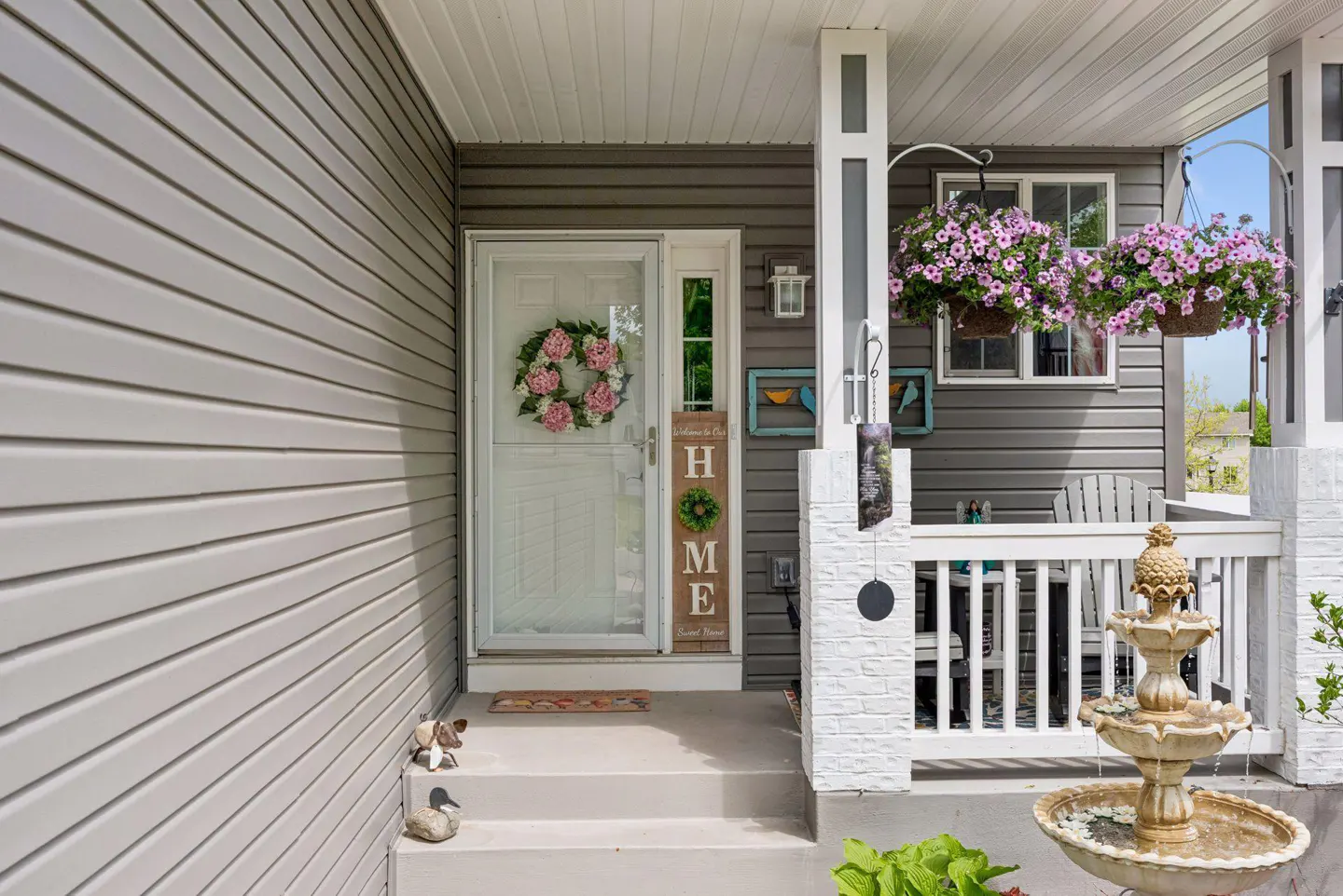 Front porch of a gray house with a white door, pink wreath, and a "HOME" sign. Hanging flower baskets and a water fountain add charm.