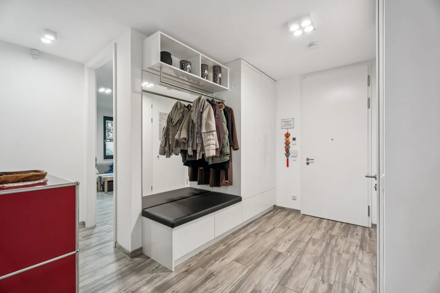 Bright entryway with wood-look floors, white walls, and a built-in bench with coat rack. Coats hang above the black cushion. A red cabinet is visible.
