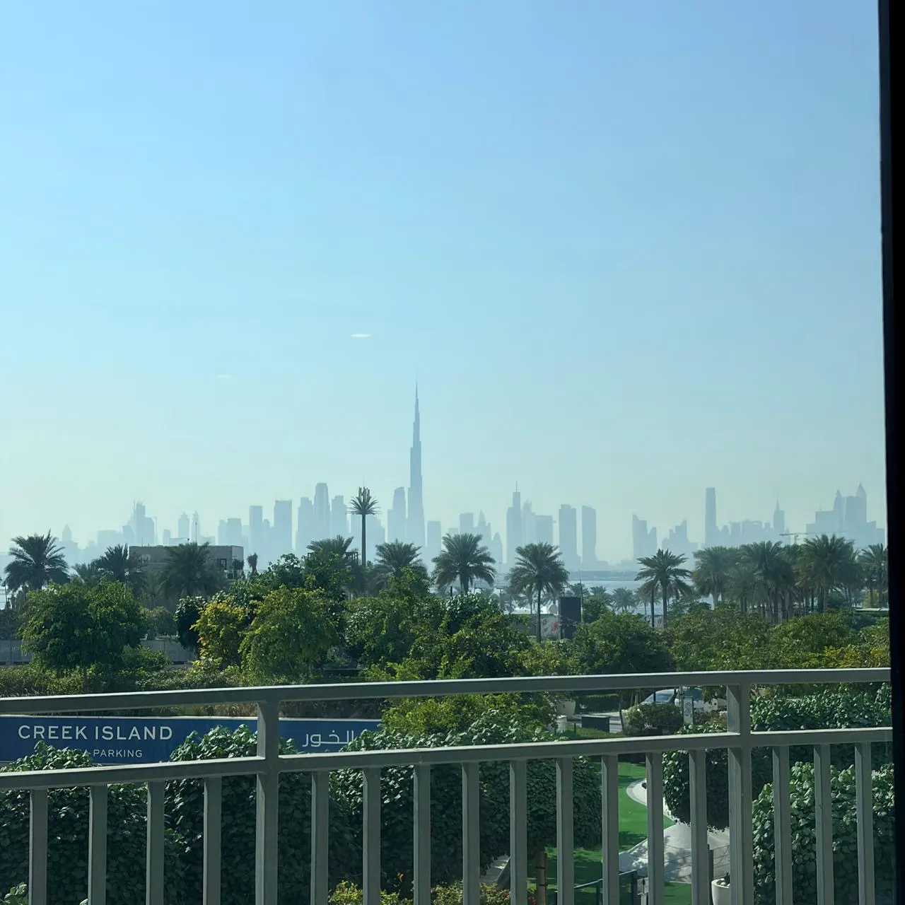 View from a balcony with white railings, overlooking green trees and the Dubai skyline under a hazy blue sky.