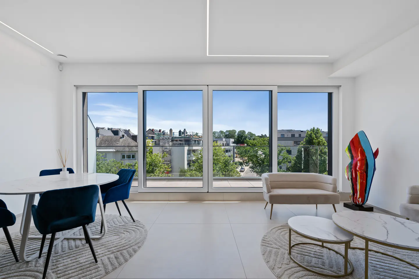 Bright, modern living room with a white table, blue chairs, and a large window overlooking a cityscape. A colorful sculpture adds a pop of art.
