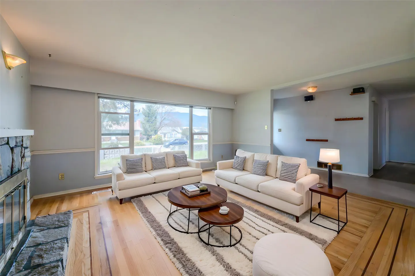 Bright living room with two white sofas, striped pillows, round wood tables, and a fireplace. A large window overlooks a green yard.