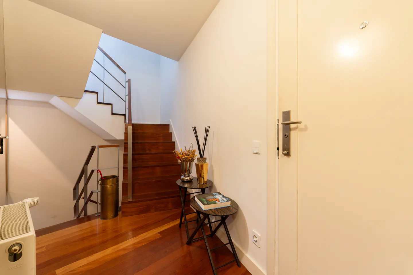 Entryway with wood floors, stairs, and a white door. Two small tables hold decor. Stairs have metal railings.