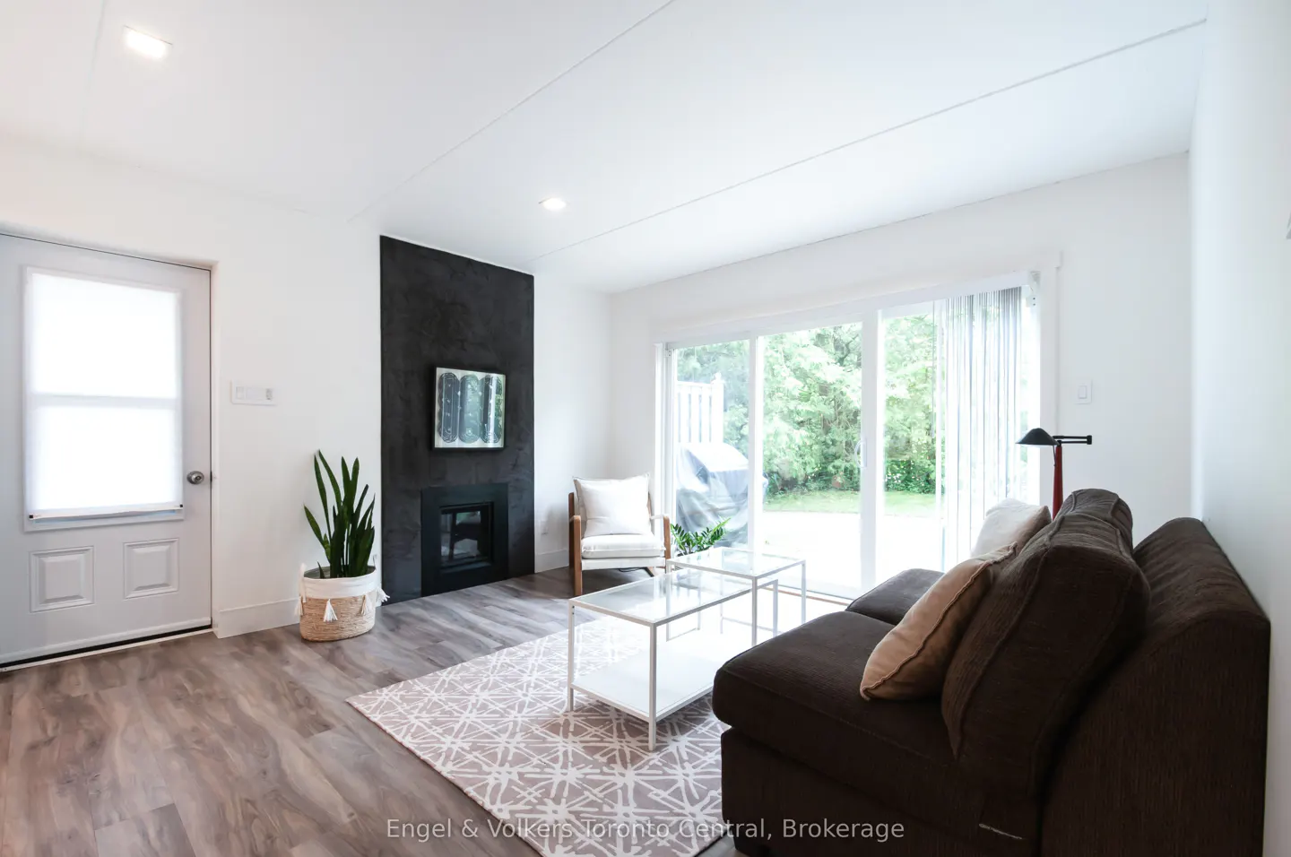 Bright living room with a brown sofa, glass tables, and a black fireplace. Sliding glass doors lead to a green backyard.