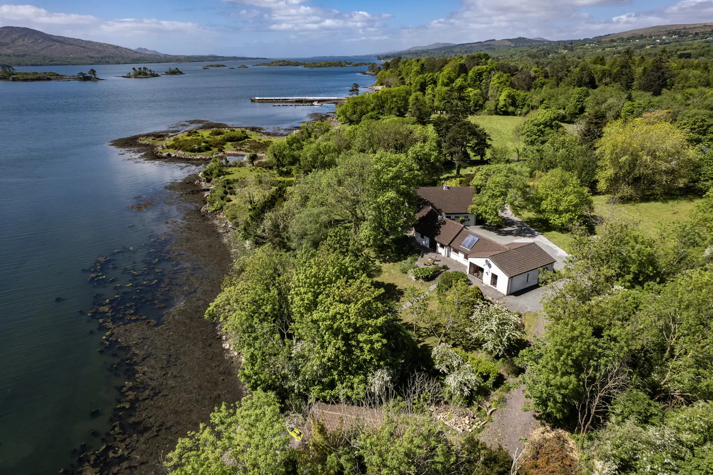 Aerial view of a white house with a brown roof, surrounded by green trees, next to a blue lake with islands.