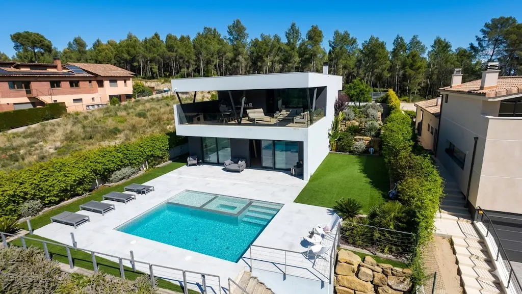 Two-story modern white house with a pool, lounge chairs, and a balcony with outdoor furniture. Trees and blue sky in the background.