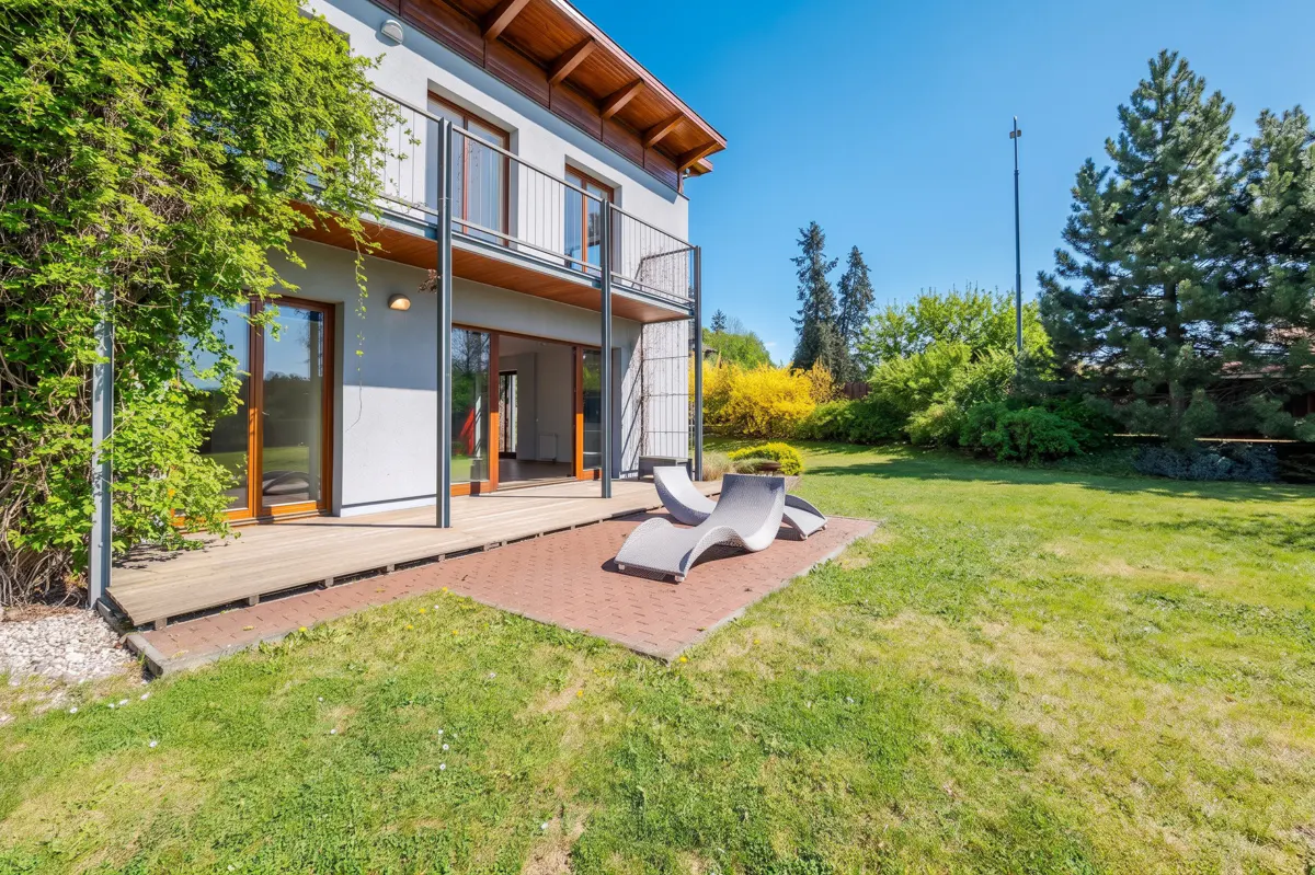 Two-story house with a balcony, wooden deck, and two gray lounge chairs on a brick patio in the backyard. Green grass and trees surround the house.