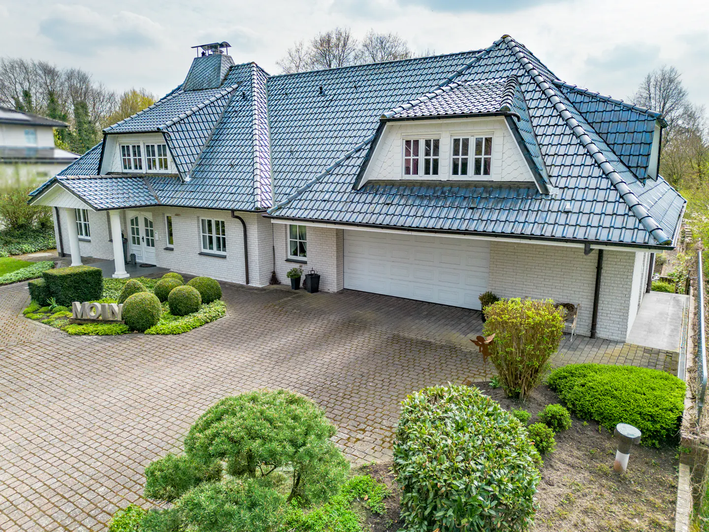 Aerial view of a white brick house with a blue tile roof and a circular driveway with green bushes.