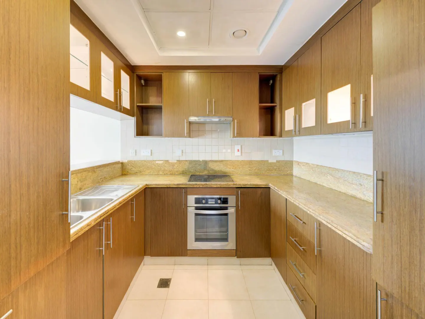 A well-lit kitchen with wood cabinets, granite countertops, stainless steel sink and oven, and white tile flooring.