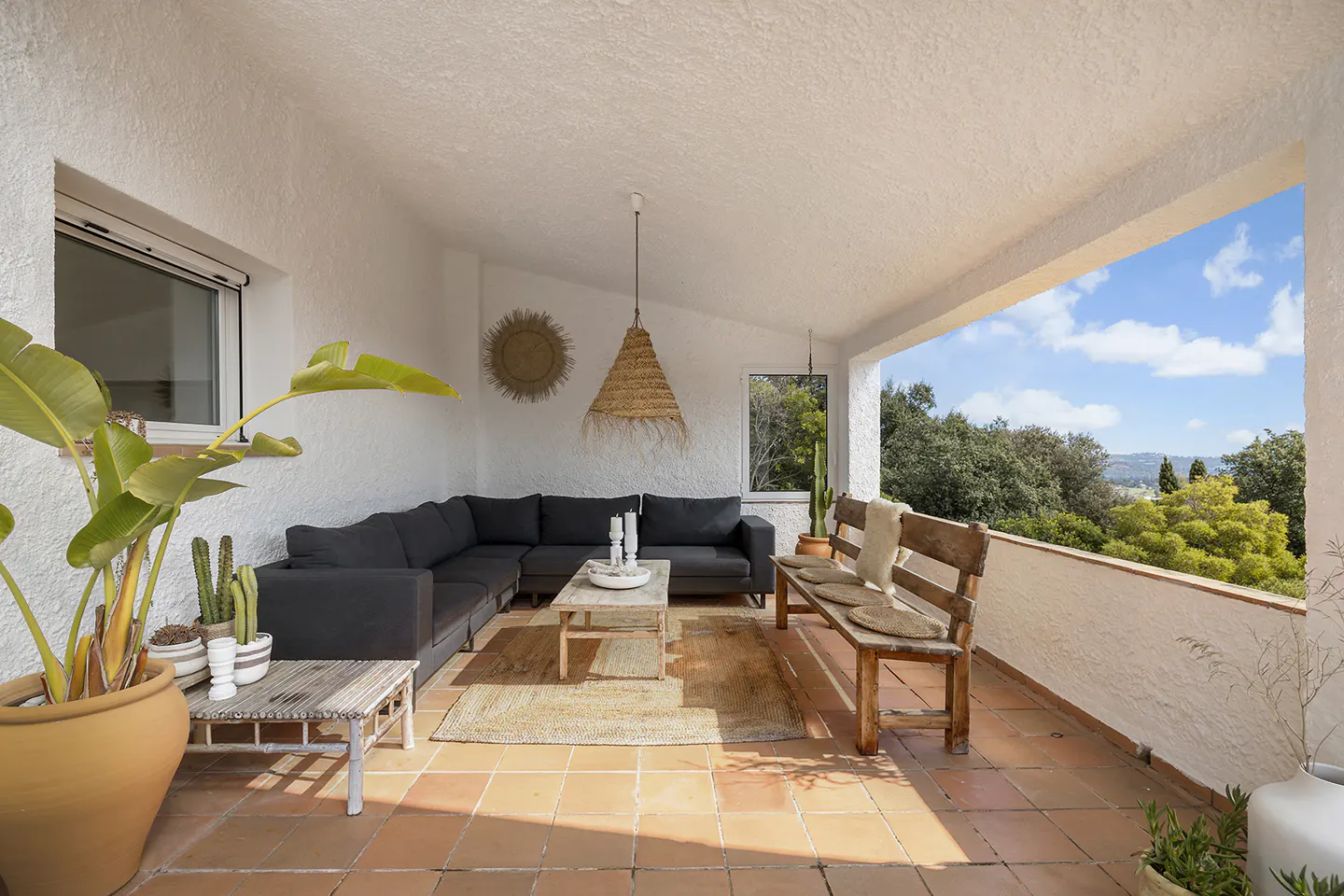 Covered patio with terracotta tile floor, white walls, and a view of trees and blue sky. A gray sectional sofa and wooden bench provide seating.