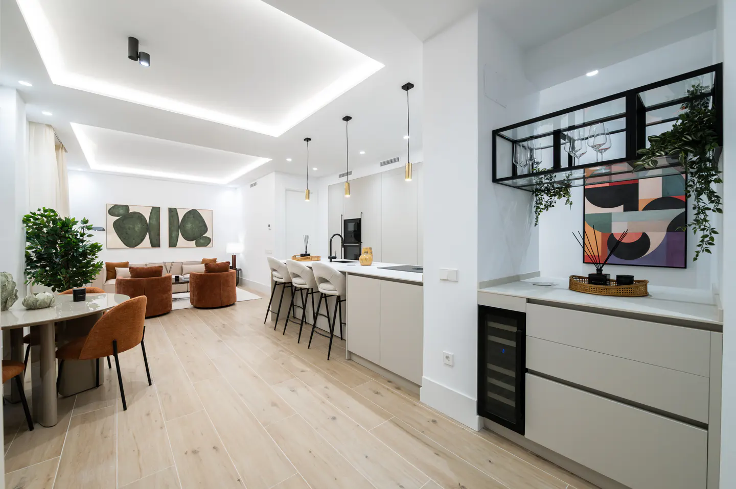 Bright, modern apartment interior with light wood floors. A dining table, kitchen island with stools, and living area with brown chairs are visible. Artwork adorns the walls.