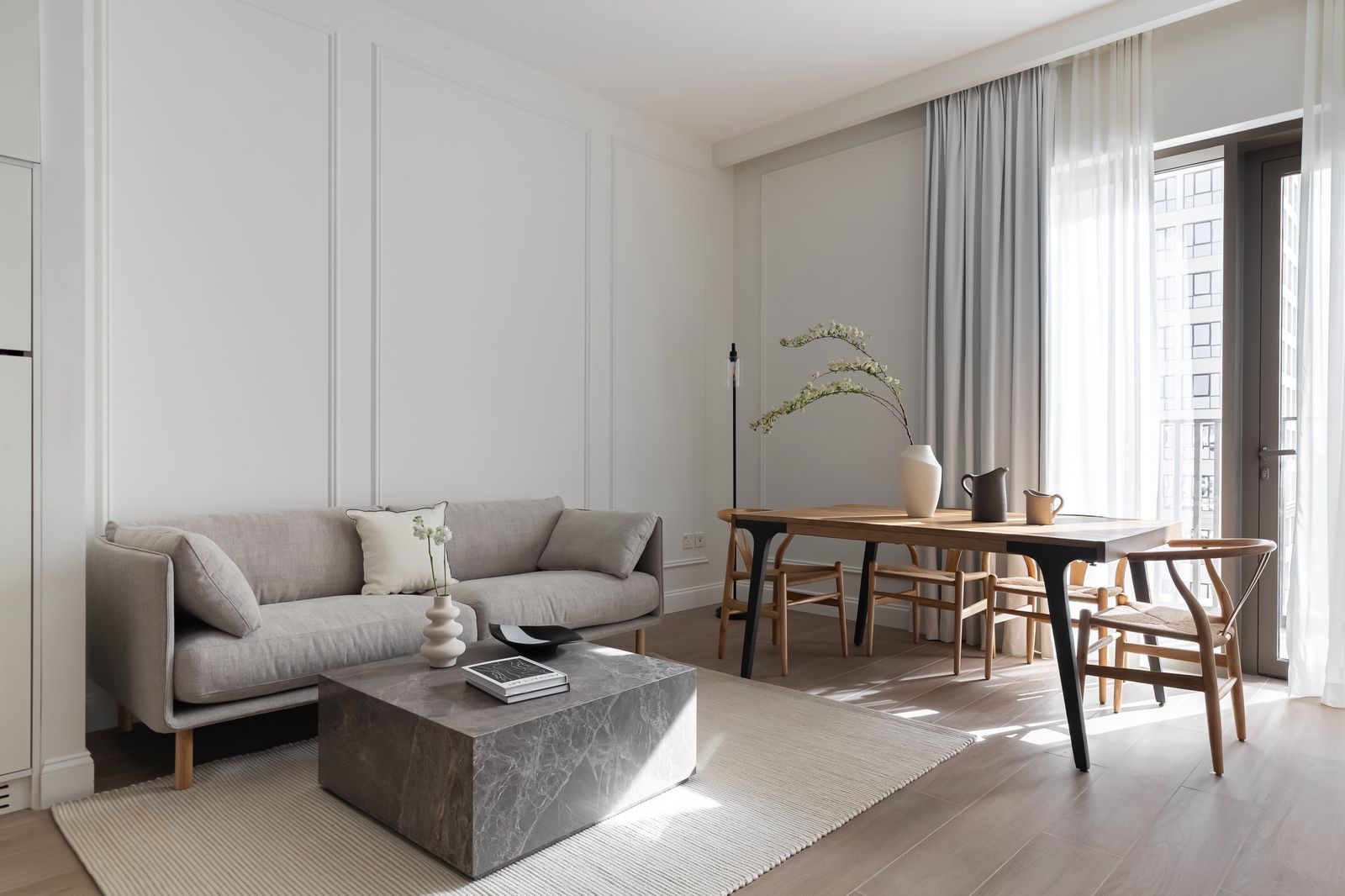 Bright living room with a gray sofa, marble coffee table, and wooden dining table with chairs near a window with gray and white curtains.