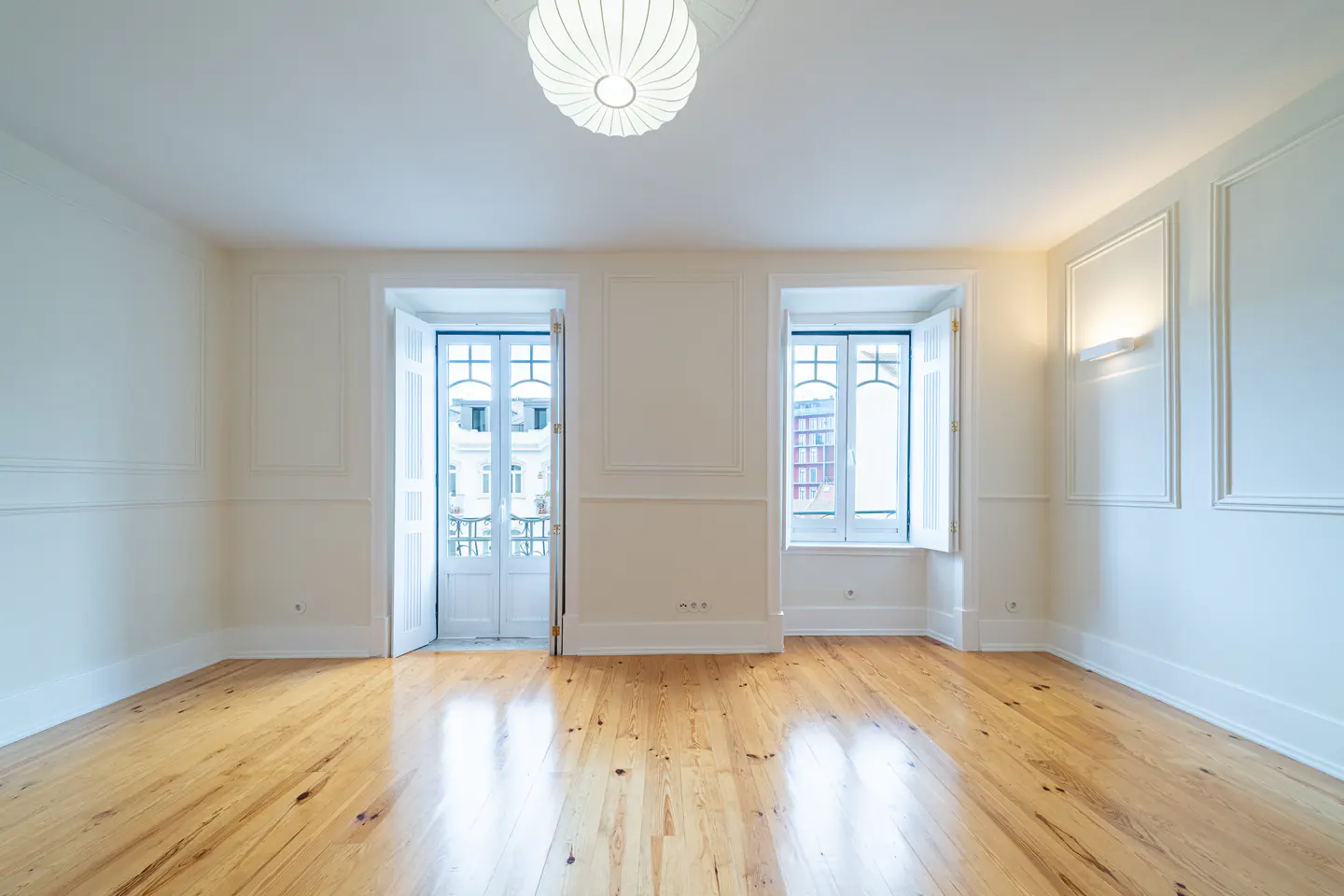 Empty room with light wood floors, white walls with molding, and two white doors/windows with views of buildings. A round light fixture hangs from the ceiling.