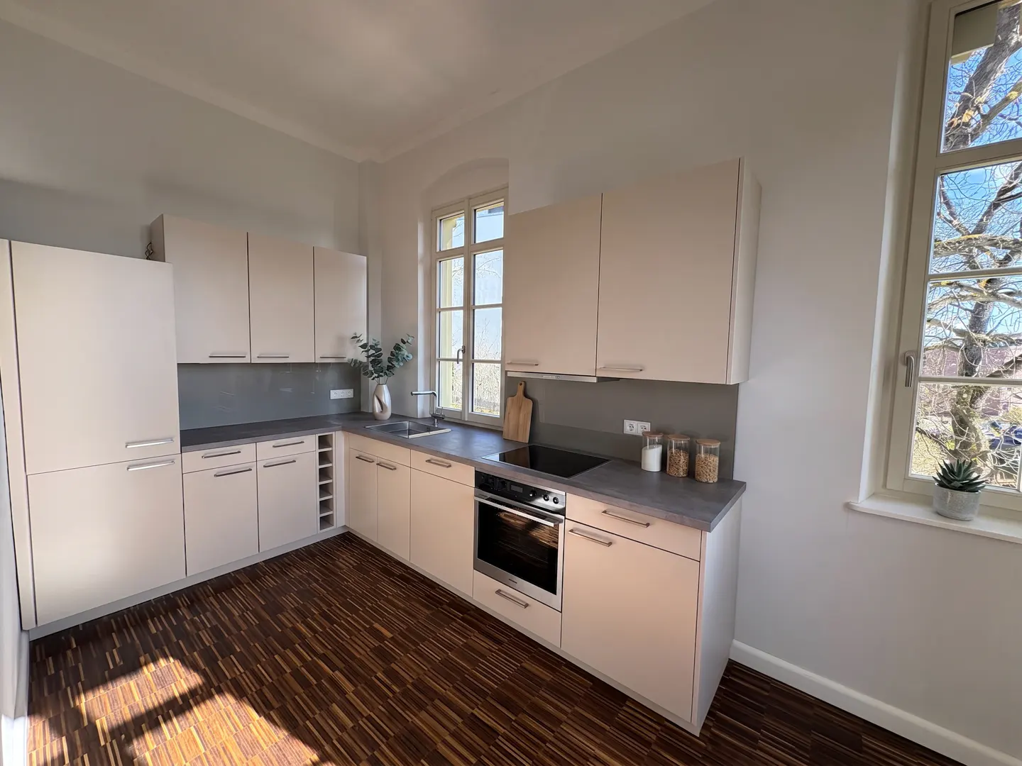 Bright kitchen with white cabinets, gray countertops, and wood floors. A window shows trees outside.