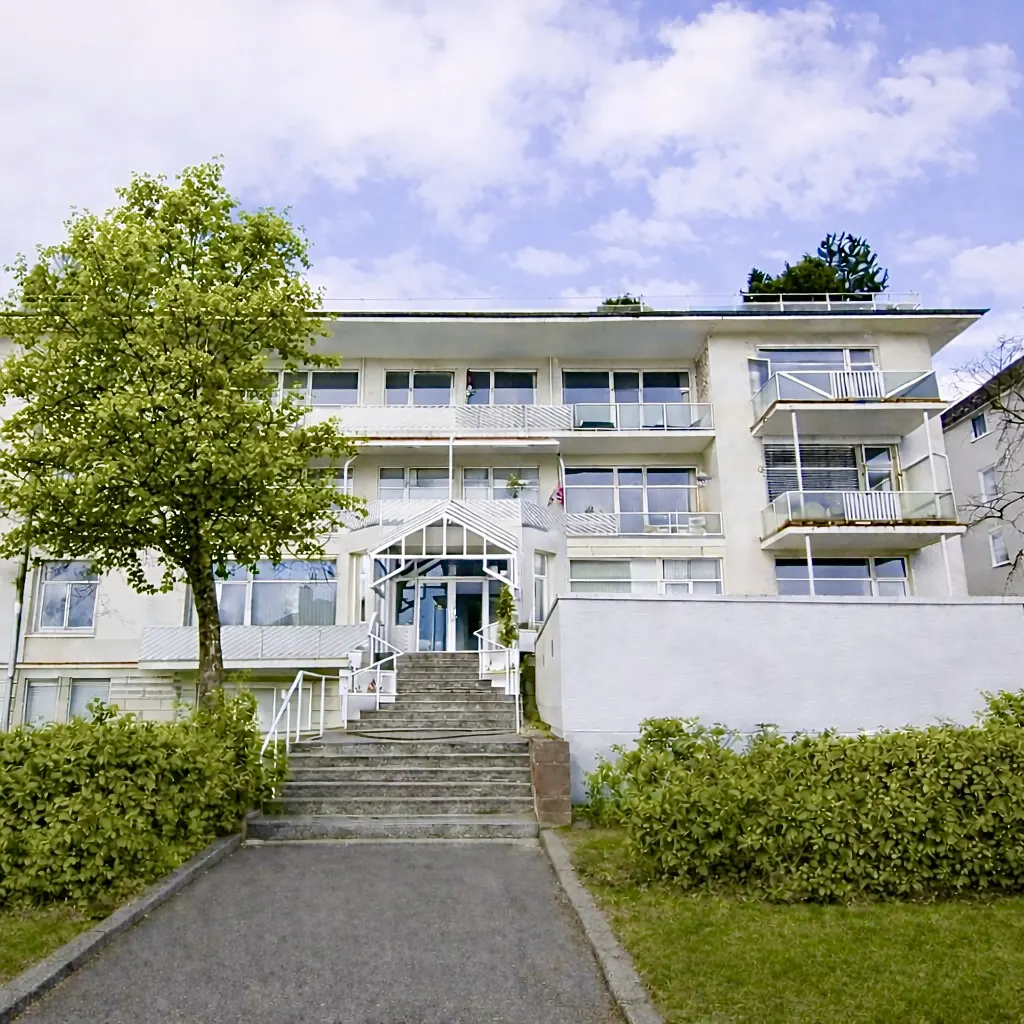 Exterior of a white, multi-story apartment building with balconies and a glass-covered entrance. A large tree stands to the left.