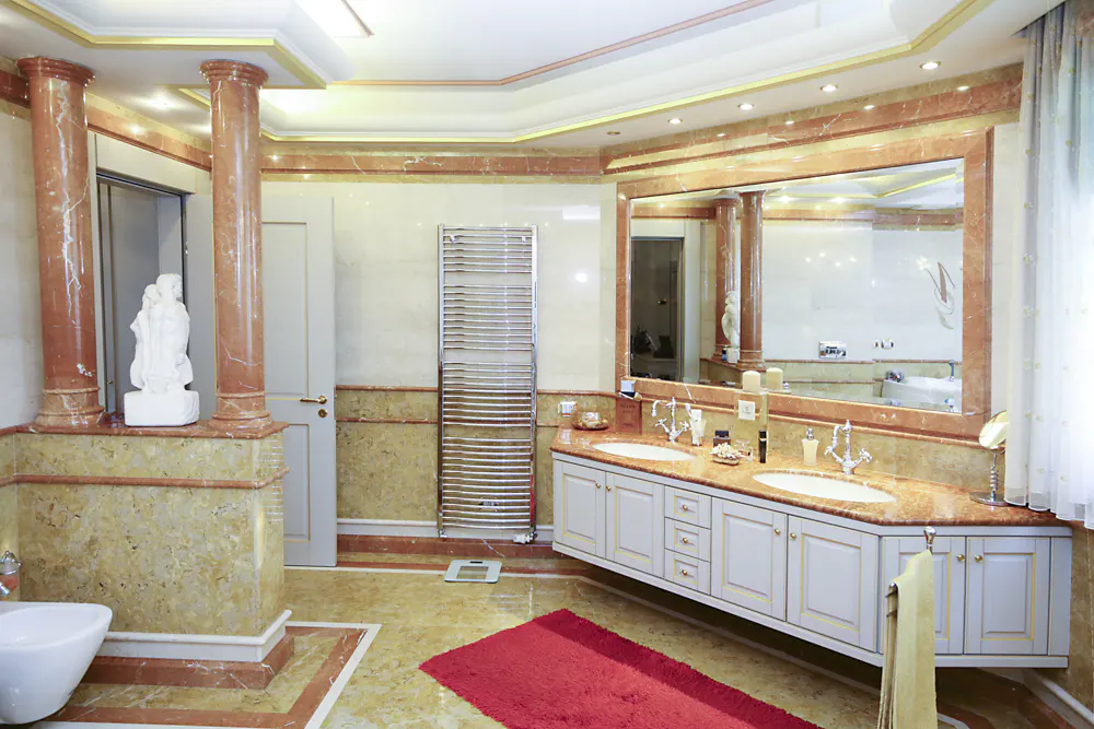 A luxurious bathroom with marble columns, a double sink vanity, and a red rug on a marble floor.