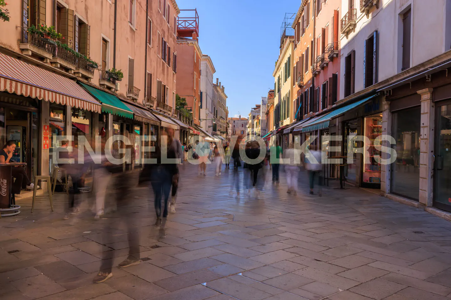 Busy Venice street with shops and people. Buildings are peach and orange with awnings. Blue sky.