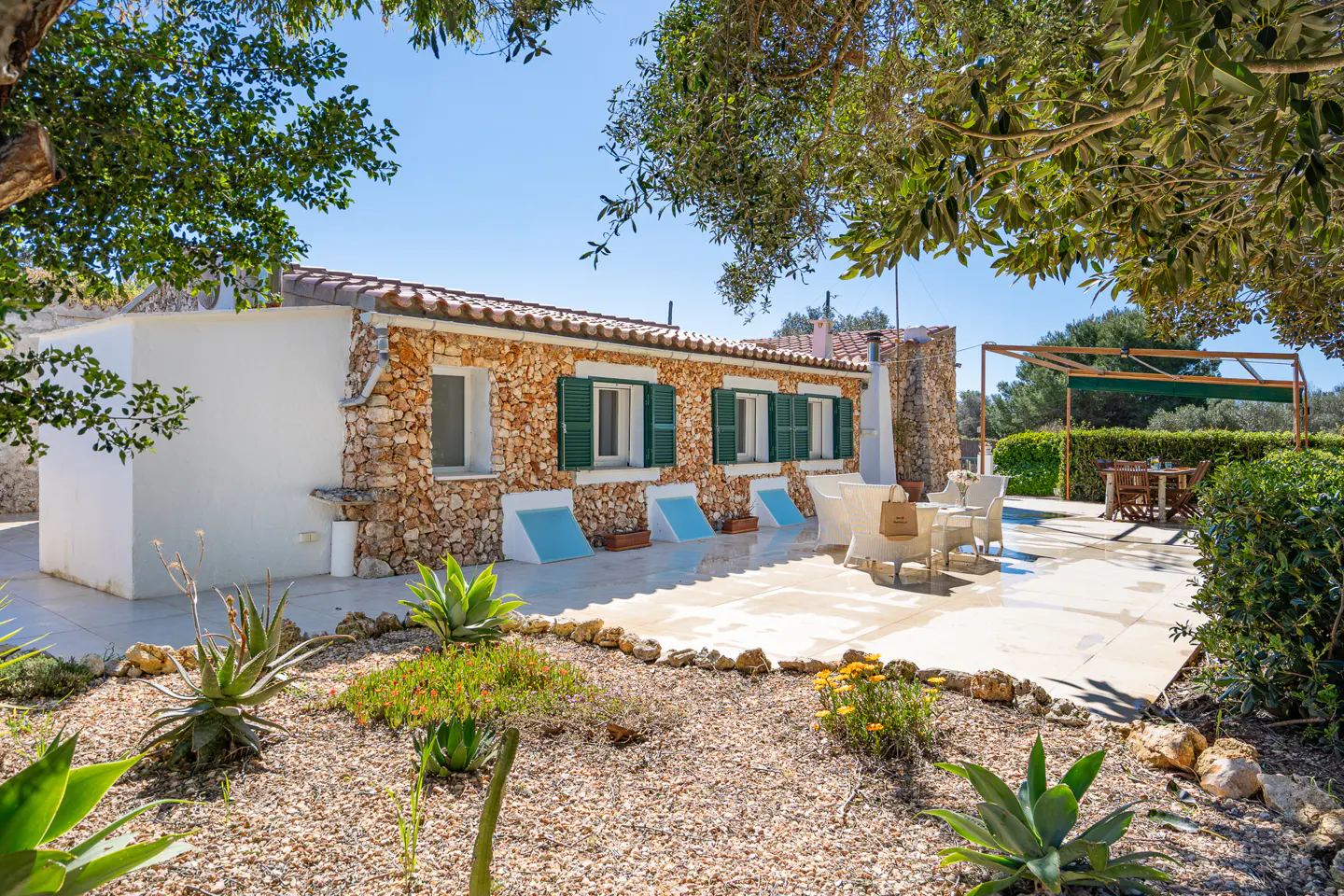 Exterior of a stone house with green shutters, a tiled roof, and a patio with outdoor furniture. A garden with gravel and plants is in the foreground.