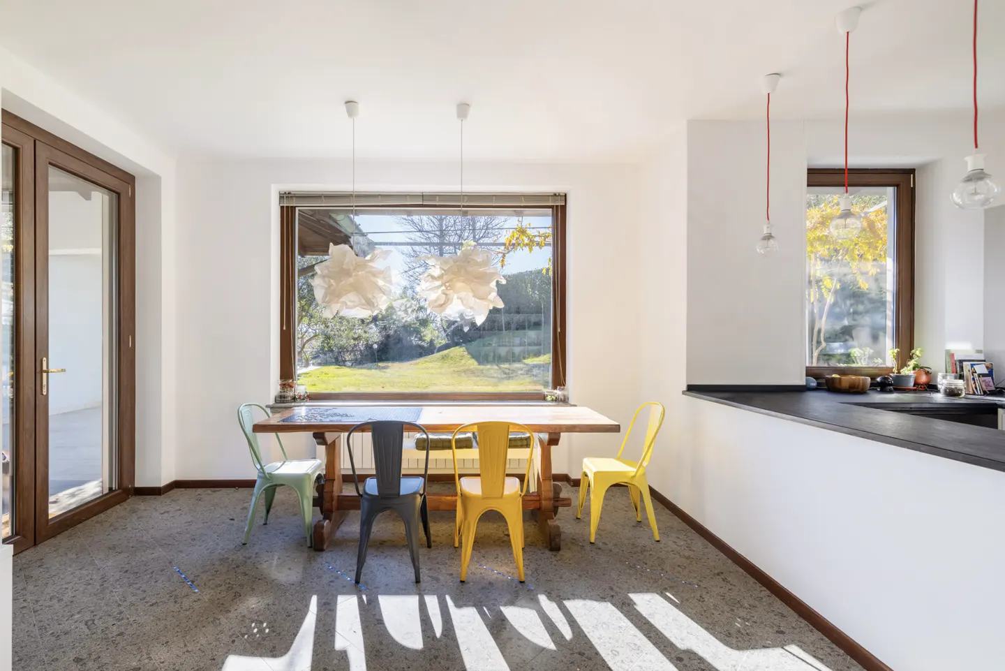 Bright dining room with a wooden table and colorful chairs under pendant lights. A large window overlooks a green yard.