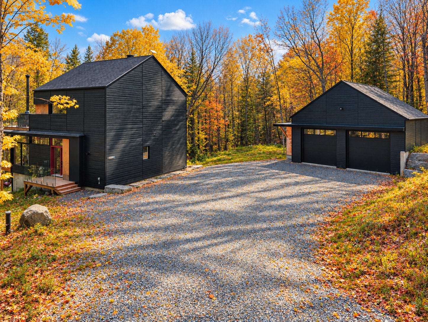 Modern black house and detached garage with a gravel driveway surrounded by vibrant autumn trees under a blue sky. [AI-generated text]