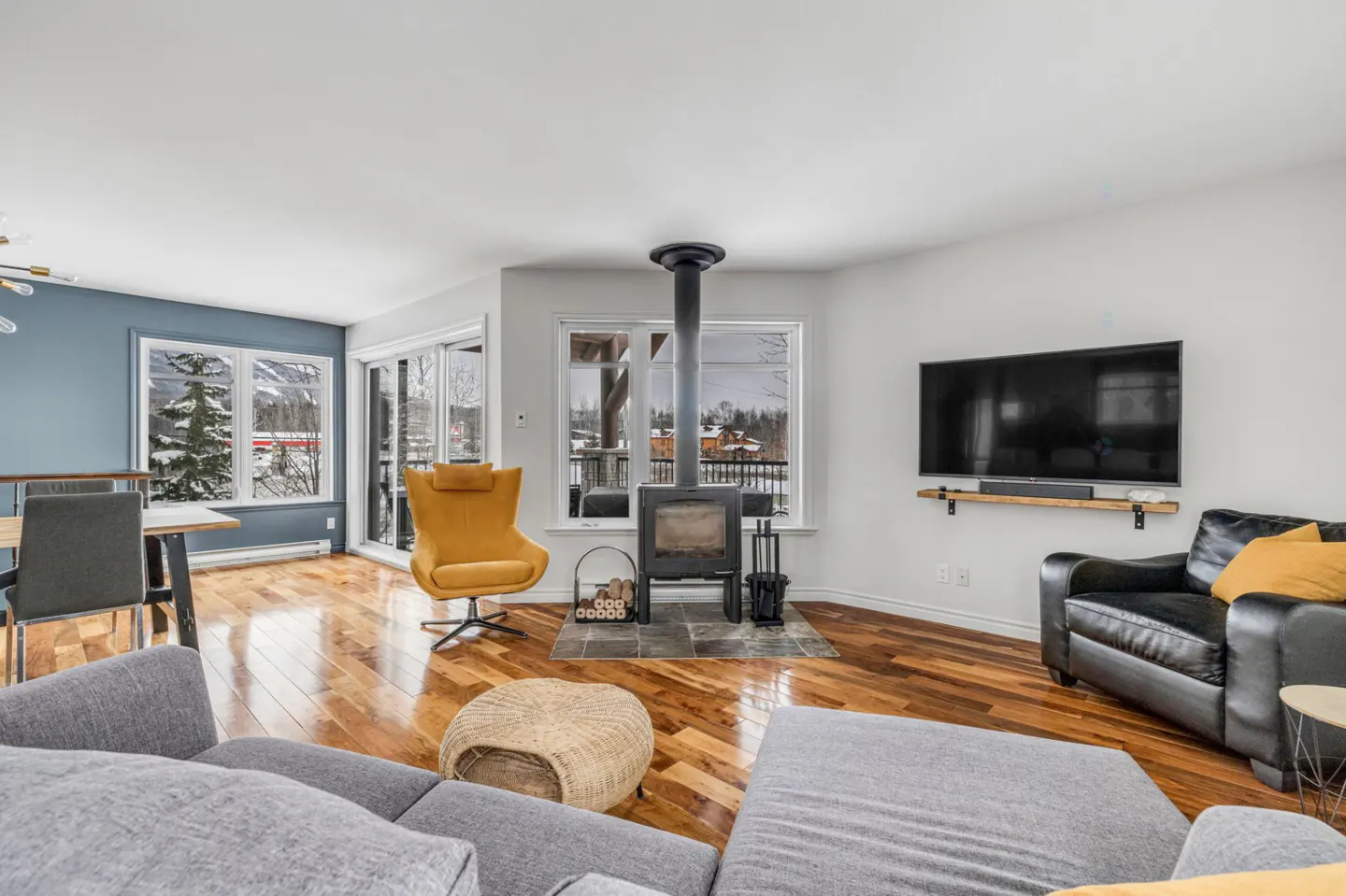 Bright living room with hardwood floors, a gray sofa, a black leather chair, and a yellow swivel chair near a wood-burning stove.
