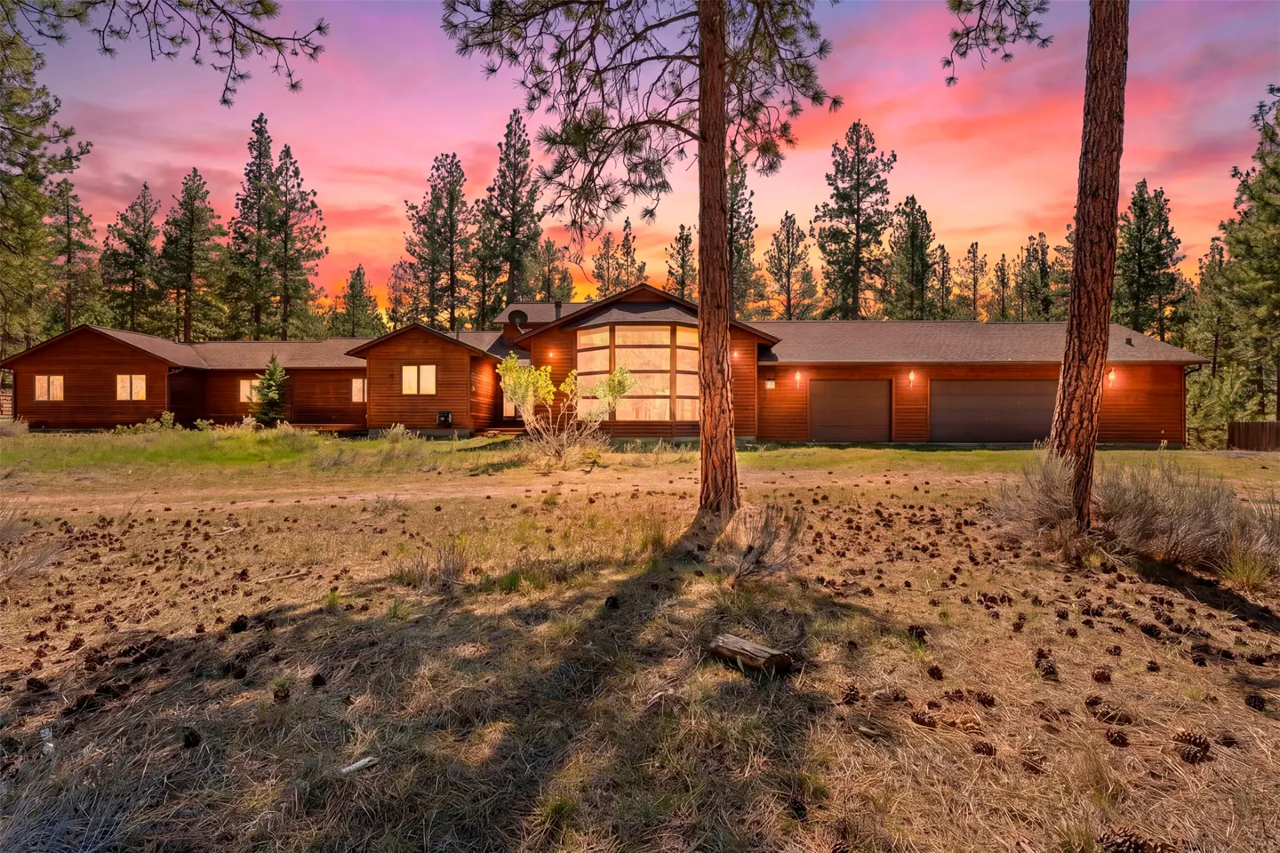 A long, brown, wood house with a two-car garage is surrounded by tall pine trees at sunset.