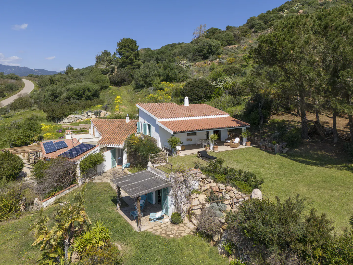 Aerial view of a white house with a red tile roof, blue shutters, and solar panels, surrounded by green trees and grass.