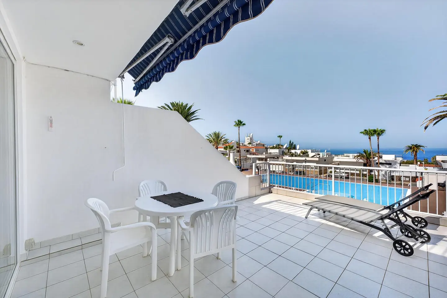 Tiled balcony with a white table, four chairs, a lounge chair, and a blue striped awning overlooking a pool and the sea.