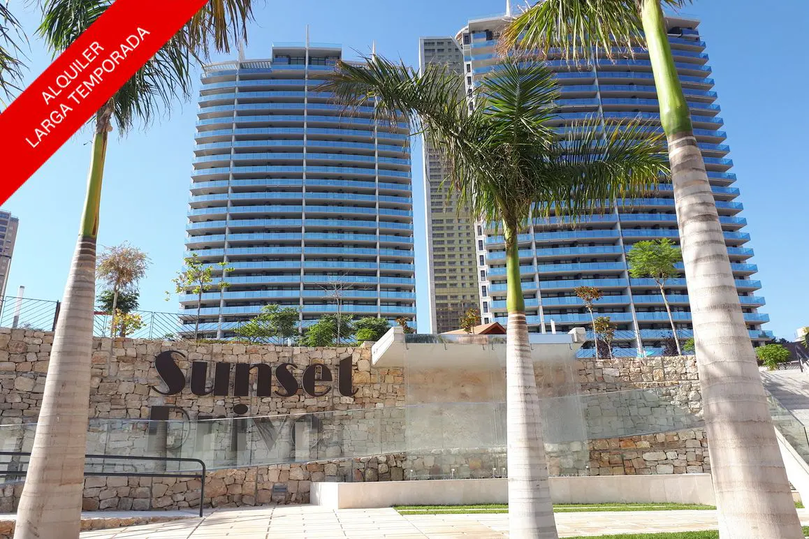 Exterior view of Sunset Drive apartments with palm trees and a stone wall sign. Blue balconies and a red "Alquiler Larga Temporada" banner are visible.
