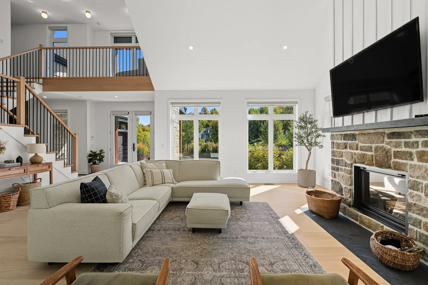 Bright, modern living room with a large sectional sofa, stone fireplace, and large windows overlooking a green landscape. Stairs lead to a second-floor balcony.