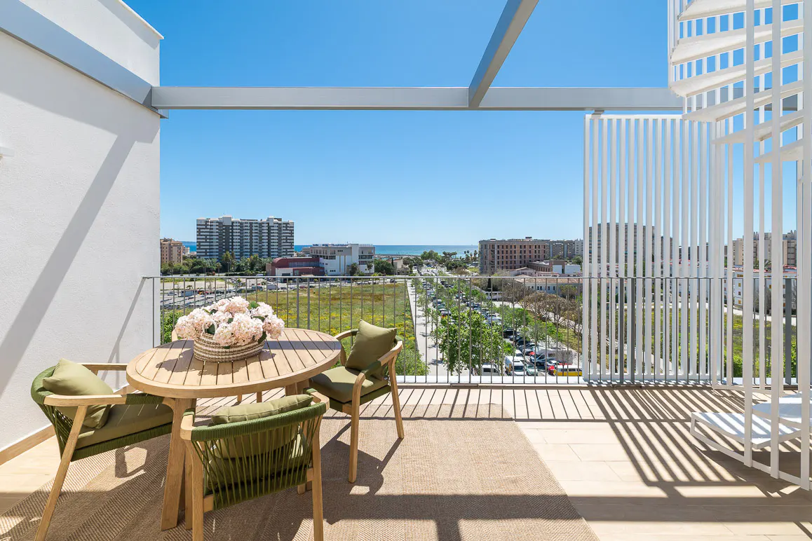 Balcony view with a round table, three green chairs, and a flower centerpiece. Cityscape and blue sky in the background.