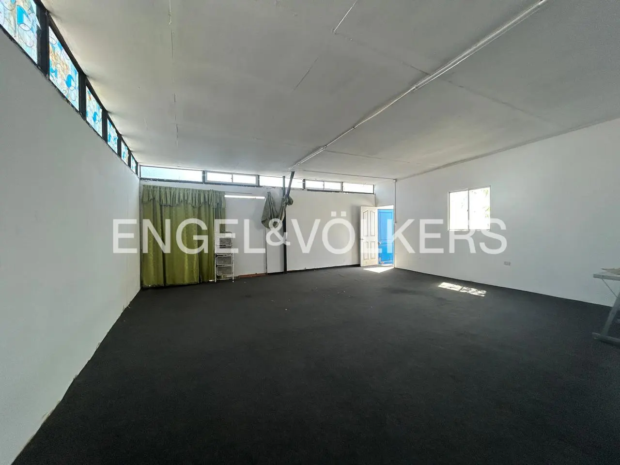 An empty room with black carpet, white walls, and a white ceiling. There are windows and a door in the background.