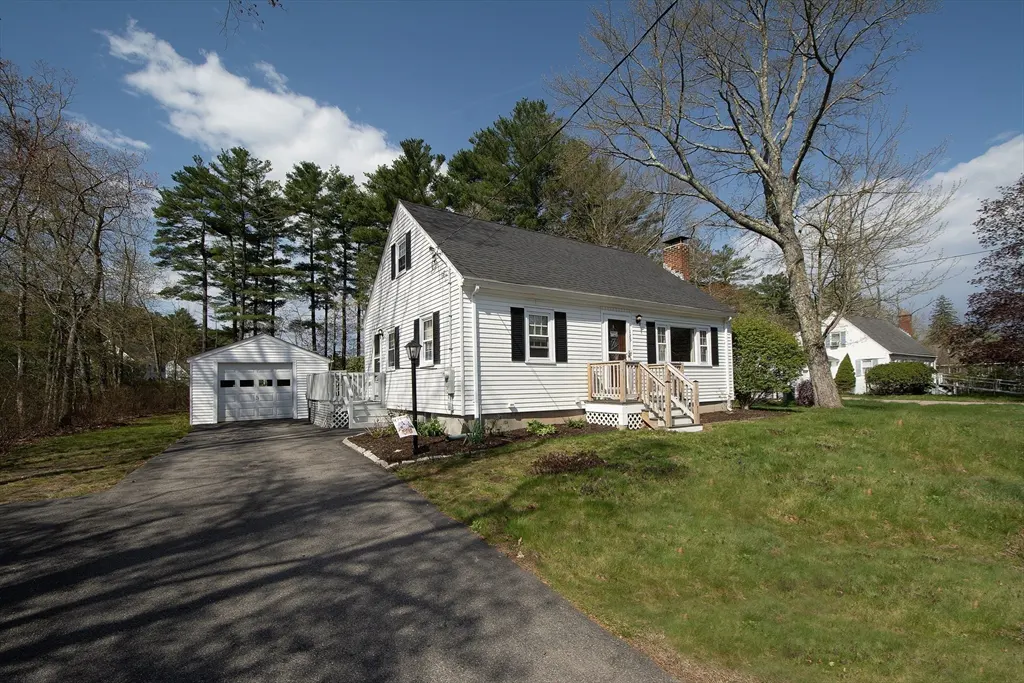 A white, one-story house with black shutters and a gray roof sits on a green lawn under a blue sky. A driveway leads to a detached garage.